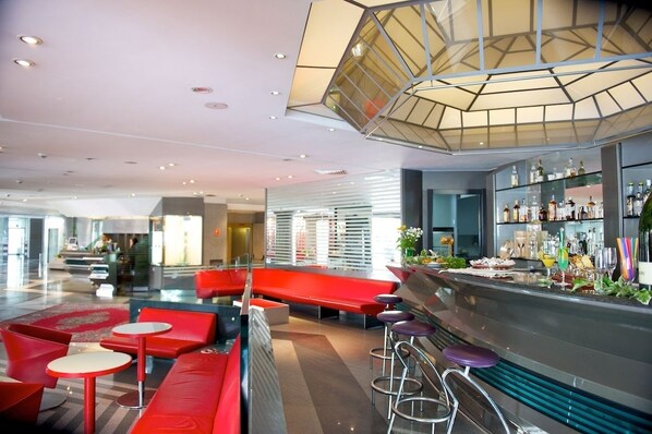 Zona de bar moderna con asientos rojos, una barra de cristal y un diseño geométrico en el techo.