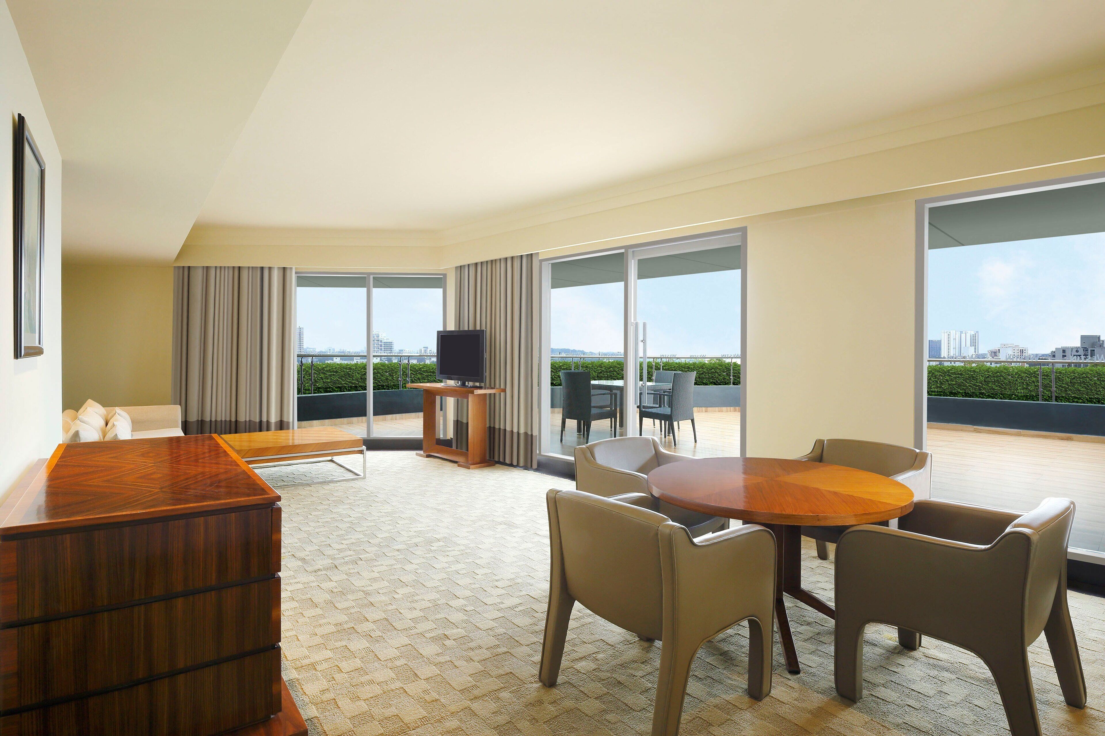 A modern hotel room with a wooden desk, a round dining table with chairs, and a balcony with a city view.