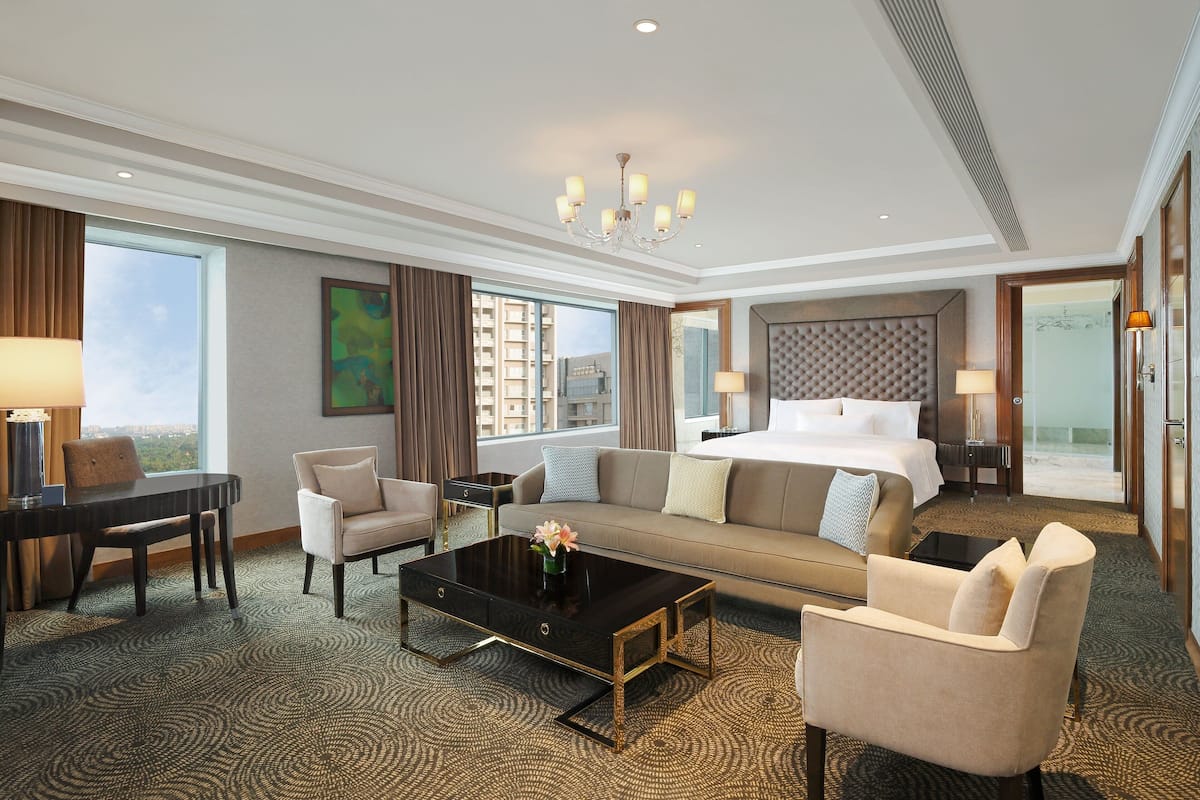 Habitación de hotel con una cama grande, un sofá, una mesa de centro, un escritorio y una silla. Se ve una ventana con vistas a edificios.