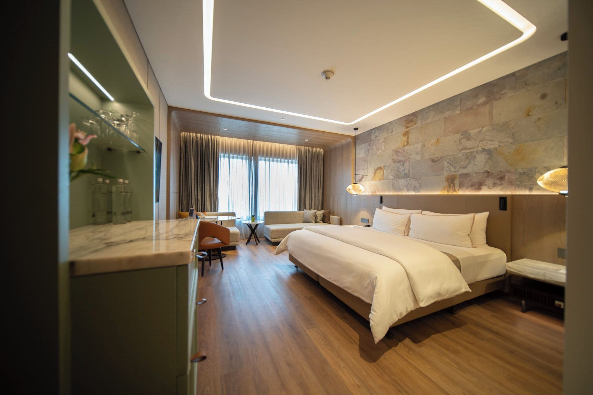Una habitación de hotel moderna con una cama grande, una zona de estar con una mesa y sillas, y vistas al exterior a través de amplios ventanales.