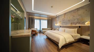 Una habitación de hotel moderna con una cama grande, una zona de estar con una mesa y sillas, y vistas al exterior a través de amplios ventanales.