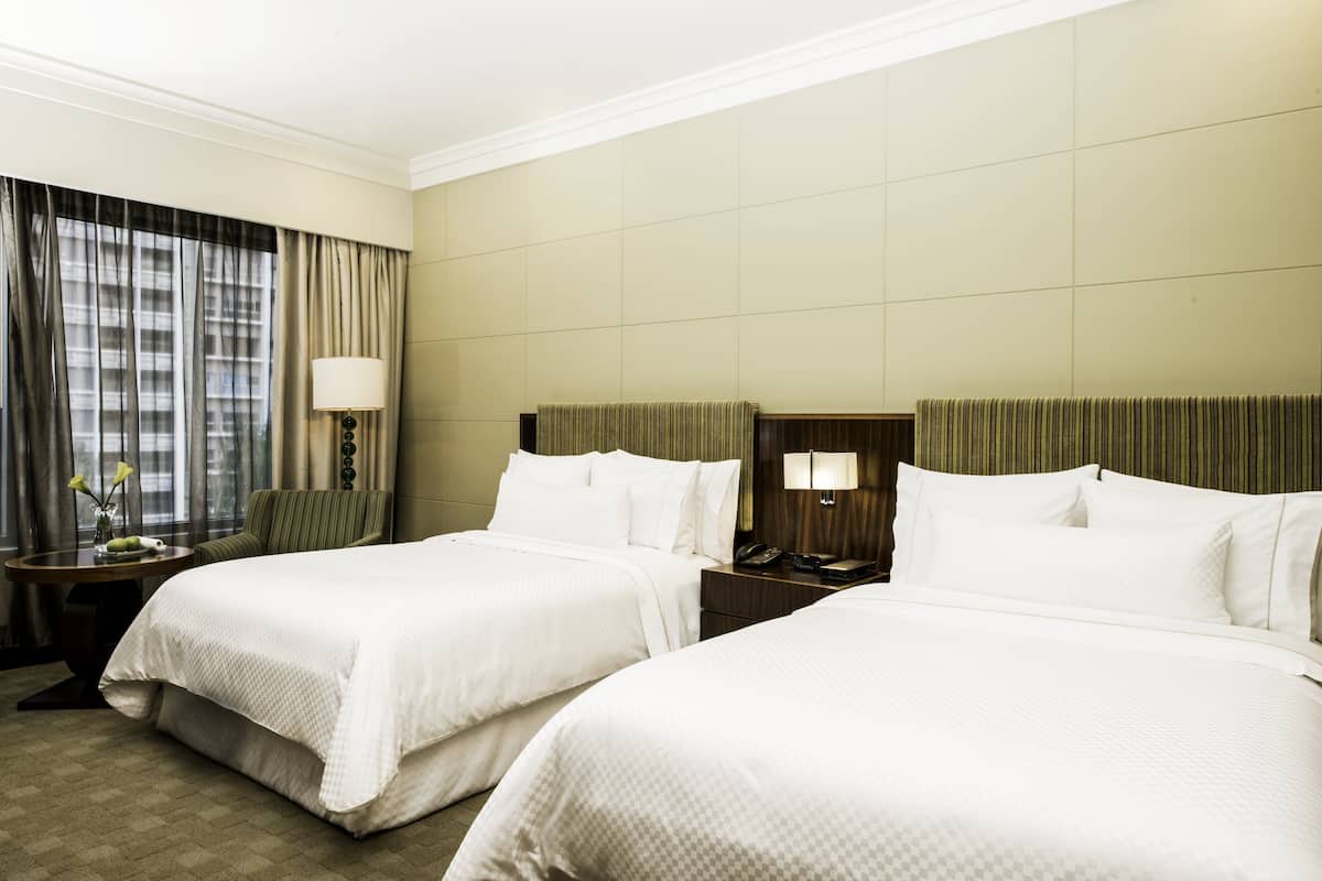Habitación de hotel con dos camas, una mesita de noche con lámpara, una silla y vista a la ciudad a través de amplias ventanas.