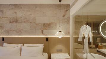 A modern hotel room with a large bed, a glass partition with a robe, and a wooden floor.