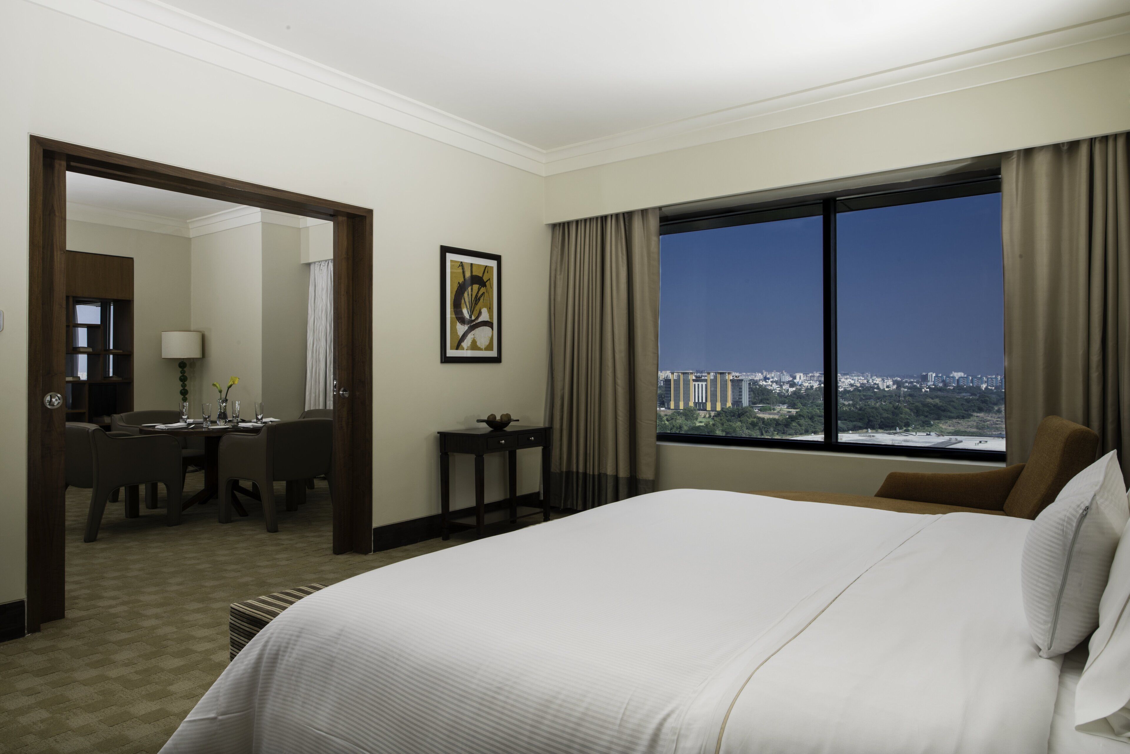 A hotel room with a large bed, a desk with a chair, a small table, a mirror, and a window with a city view.