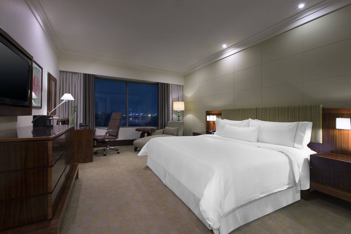 Habitación de hotel con una cama grande, un escritorio con computadora, una silla, un televisor y una ventana con cortinas.