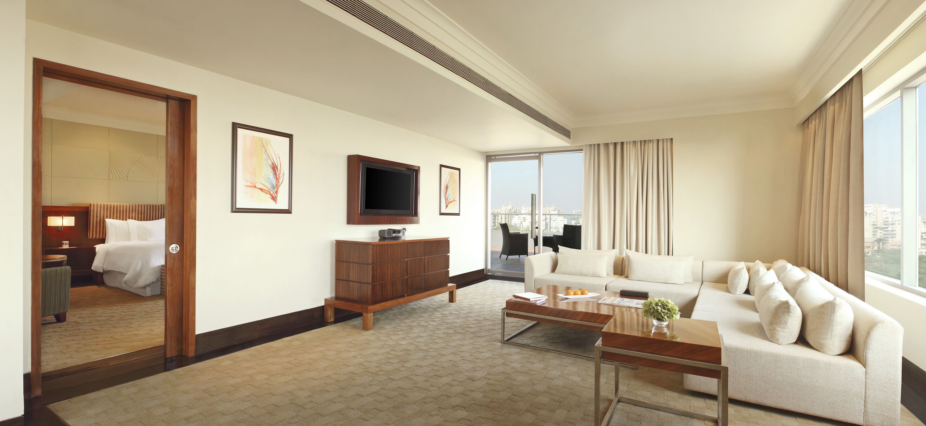 Habitación de hotel con un ventanal grande, un sofá, un televisor, un armario de madera y una cama con vistas a la ciudad.