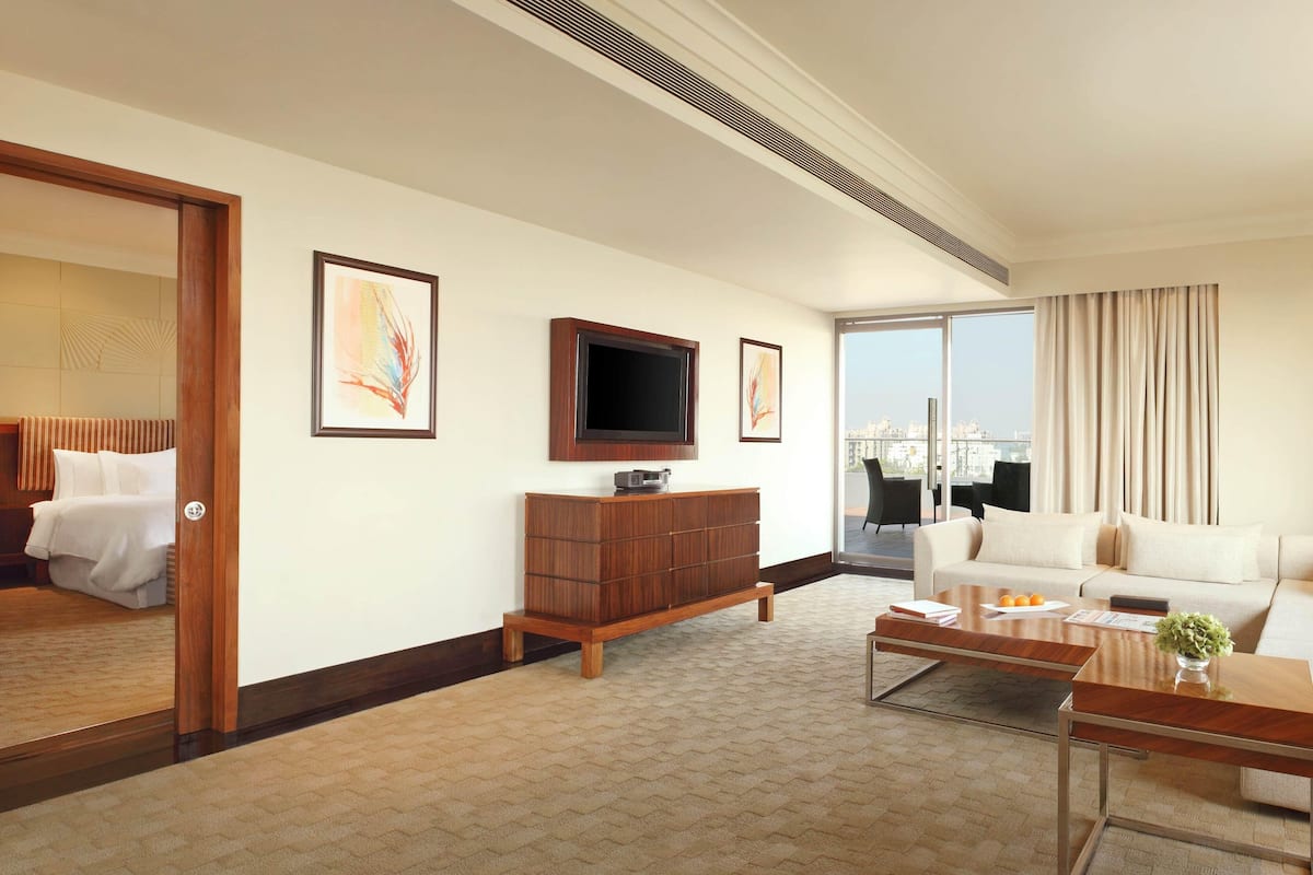 Habitación de hotel con una cama grande, un sofá, un televisor, un armario de madera y vistas a la ciudad a través de amplios ventanales.