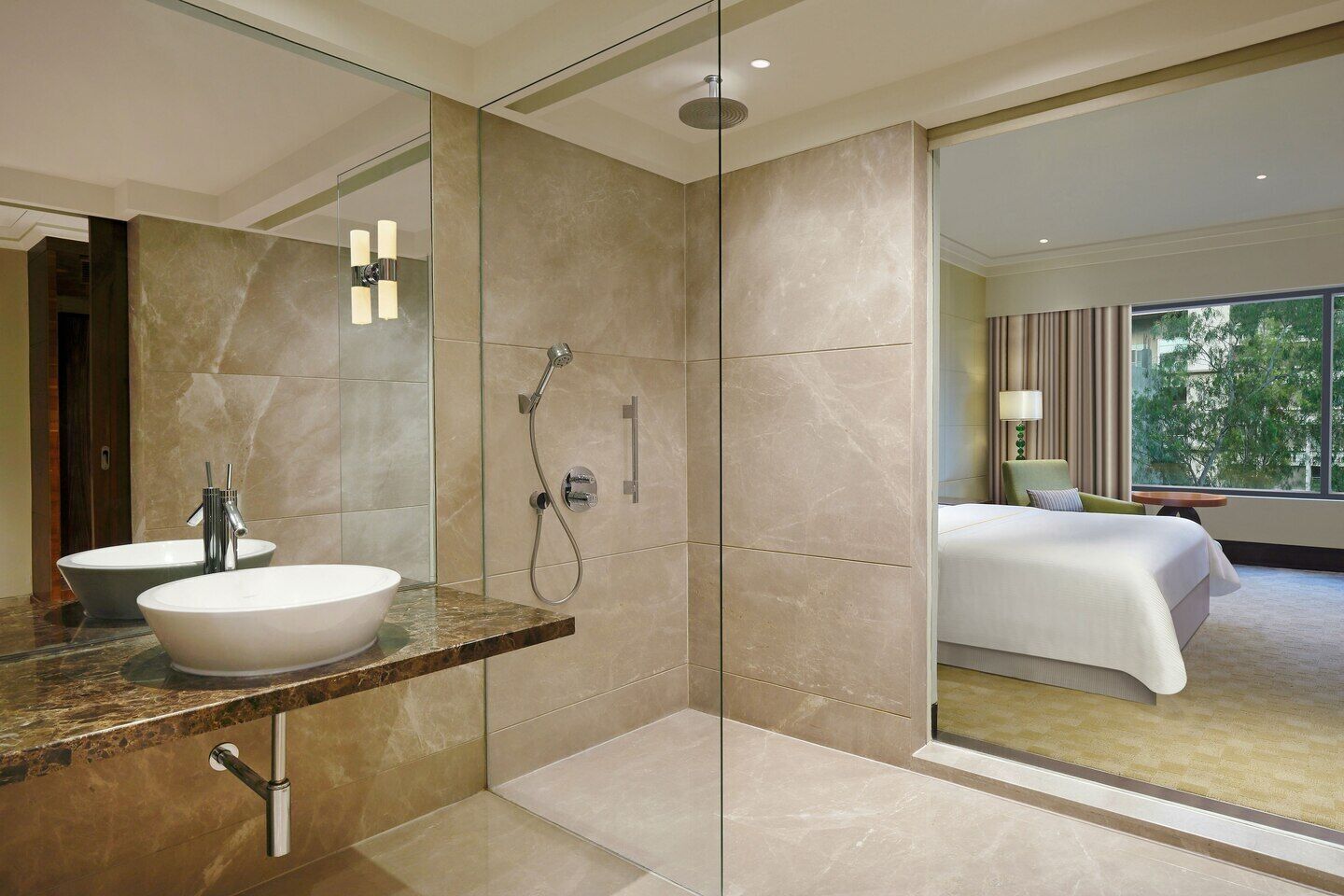 Un moderno baño de hotel con un espejo grande, un lavamanos blanco, una ducha y una cama visible en una habitación contigua.