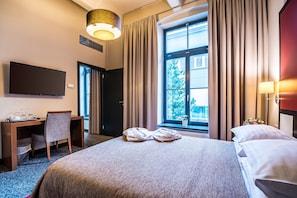 A hotel room with a large bed, a desk with a chair, a flat-screen TV, and a window with curtains.