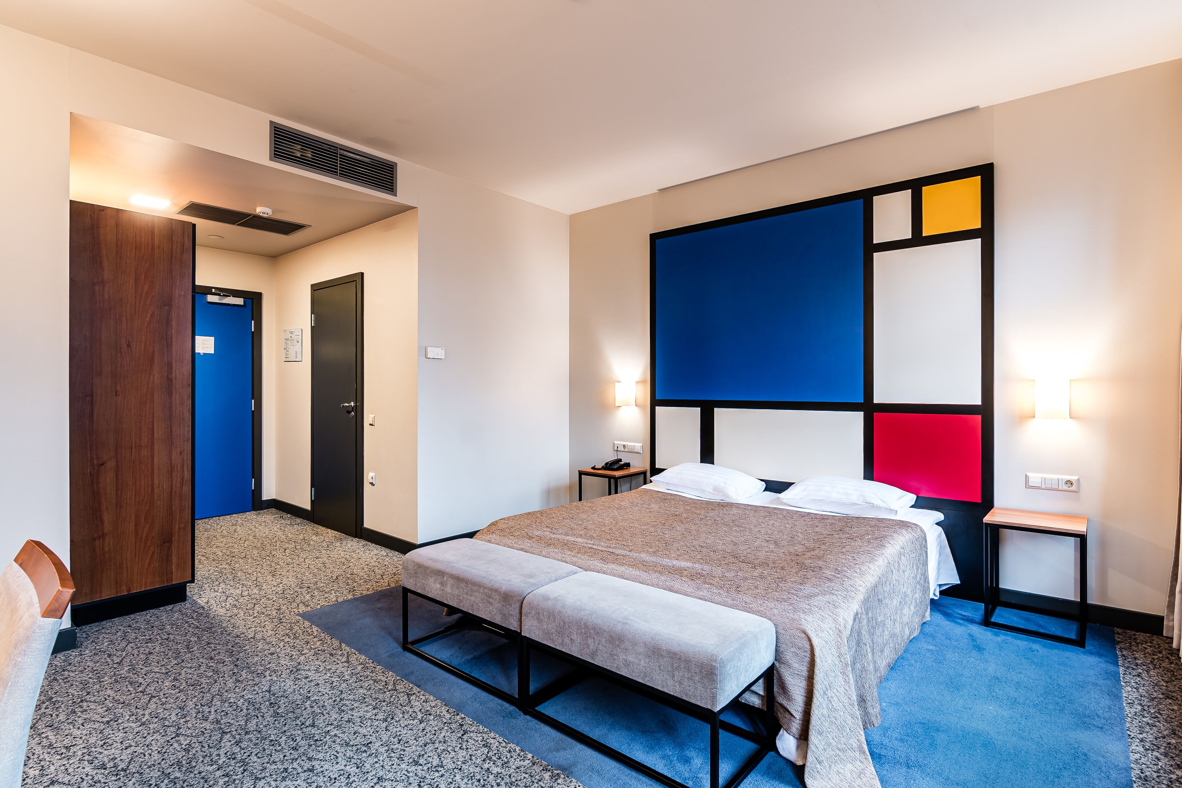 Une chambre d’hôtel avec un lit, une banquette, une table de chevet et un mur orné de motifs géométriques.