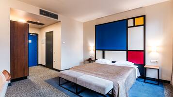 Une chambre d’hôtel avec un lit, une banquette, une table de chevet et un mur orné de motifs géométriques.