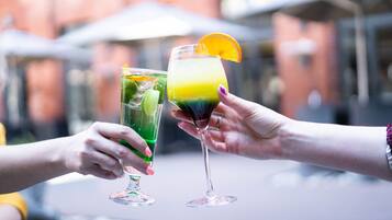 Deux personnes, chacune tenant un verre, sont assises à une table à l’extérieur, avec des bâtiments flous en arrière-plan.