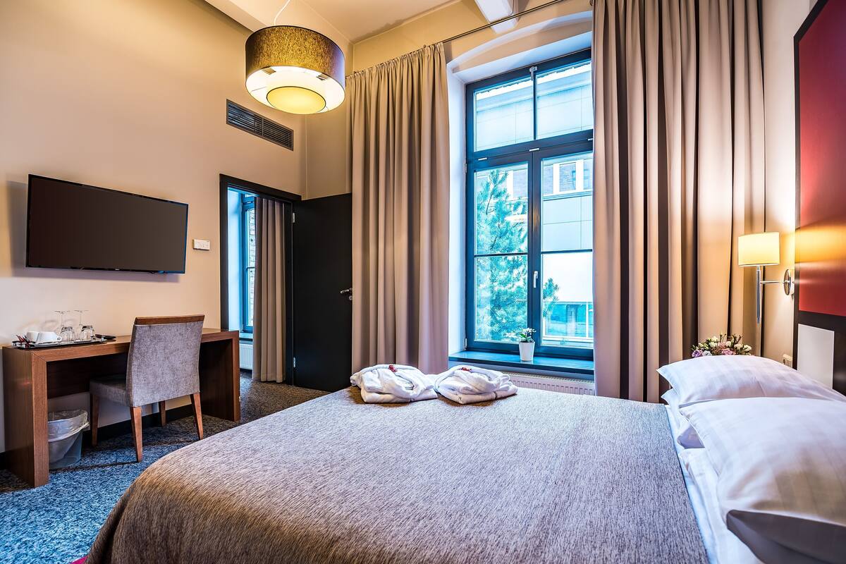 A hotel room with a bed, a desk, a chair, a TV, and a window with curtains.