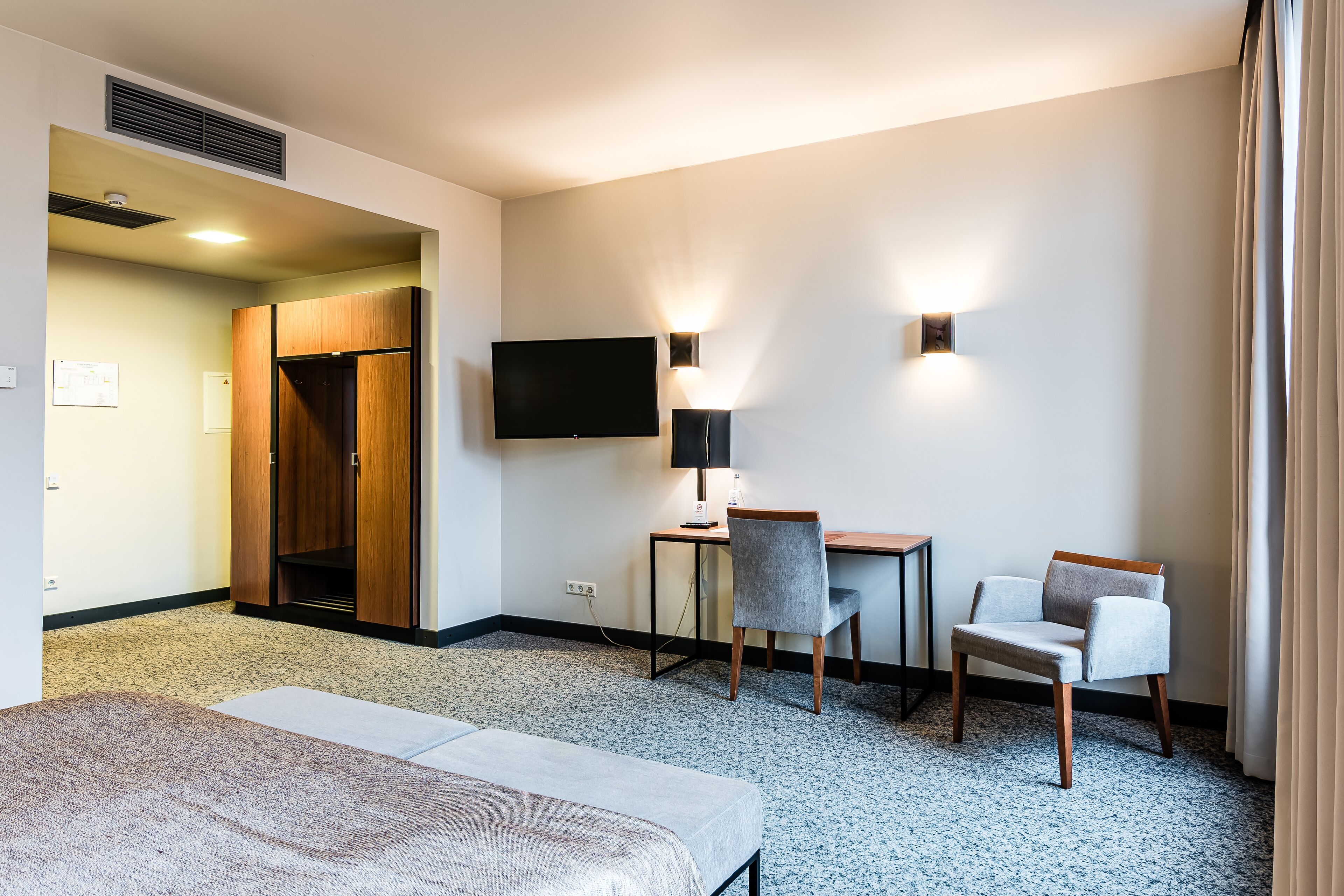 A hotel room with a bed, a desk with a chair, a television, and a wardrobe.