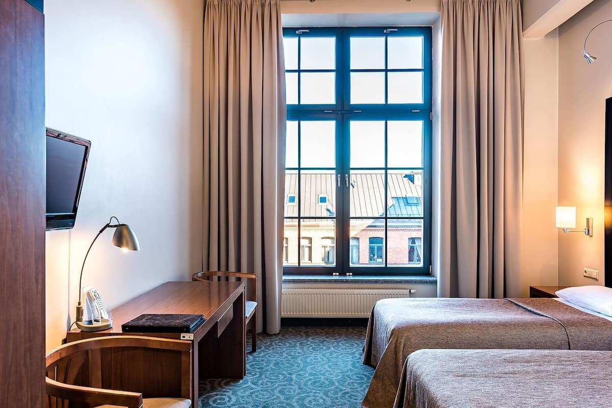 A hotel room with a large window, a desk with a lamp, two beds, and a TV on the wall.