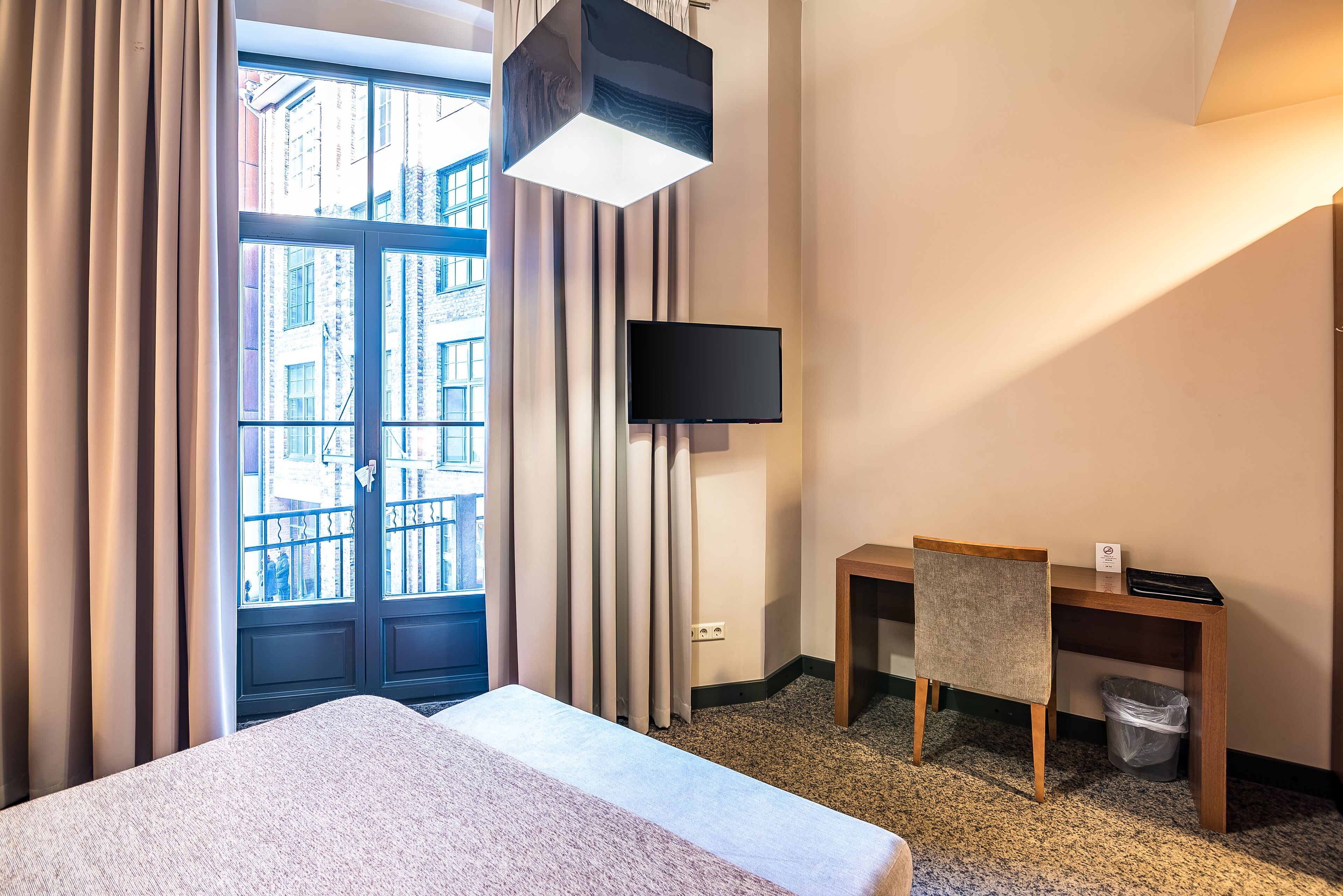 A hotel room with a bed, a desk with a chair, a television, and a view of buildings through the window.