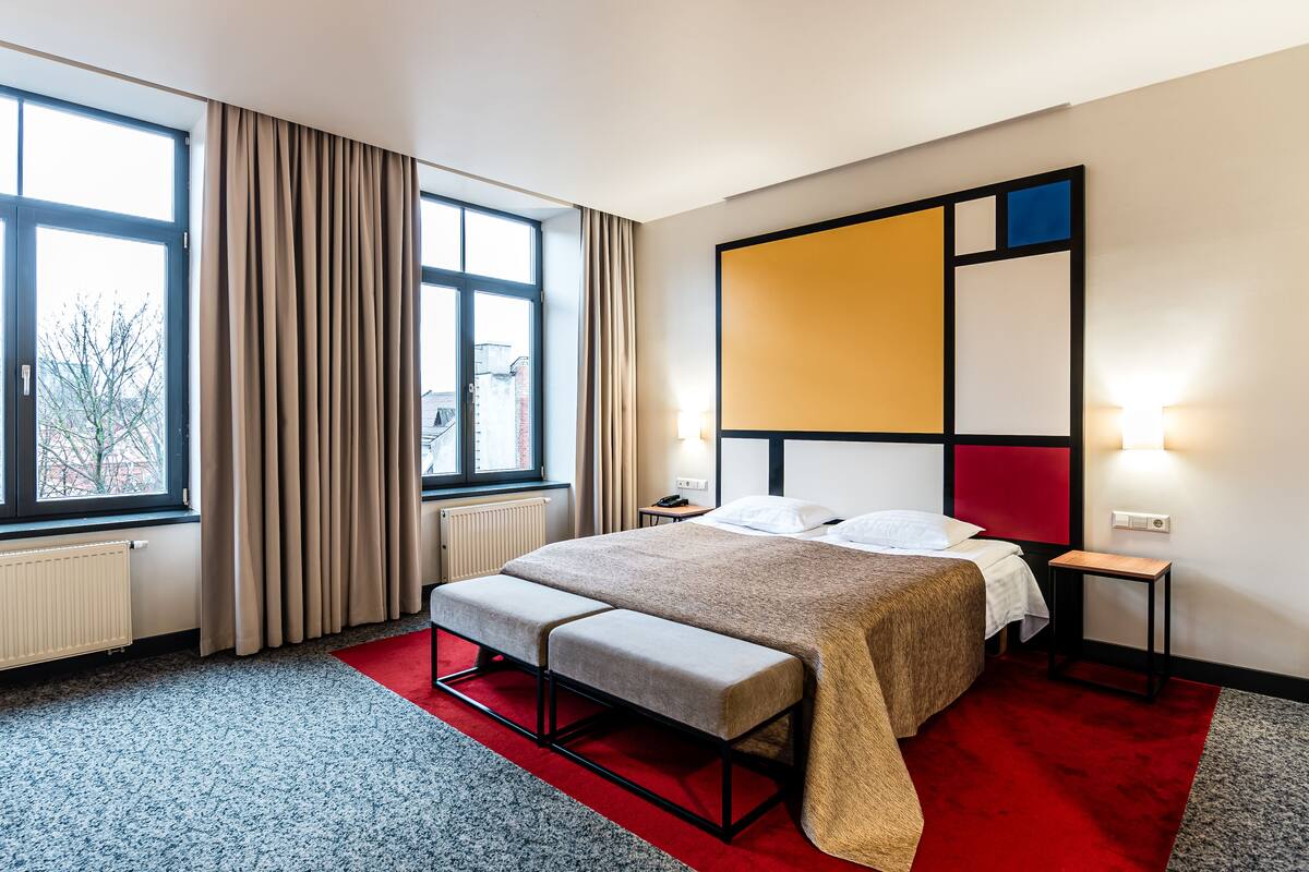A modern hotel room with a large bed, a bench, a red carpet, and a view of the city through large windows.