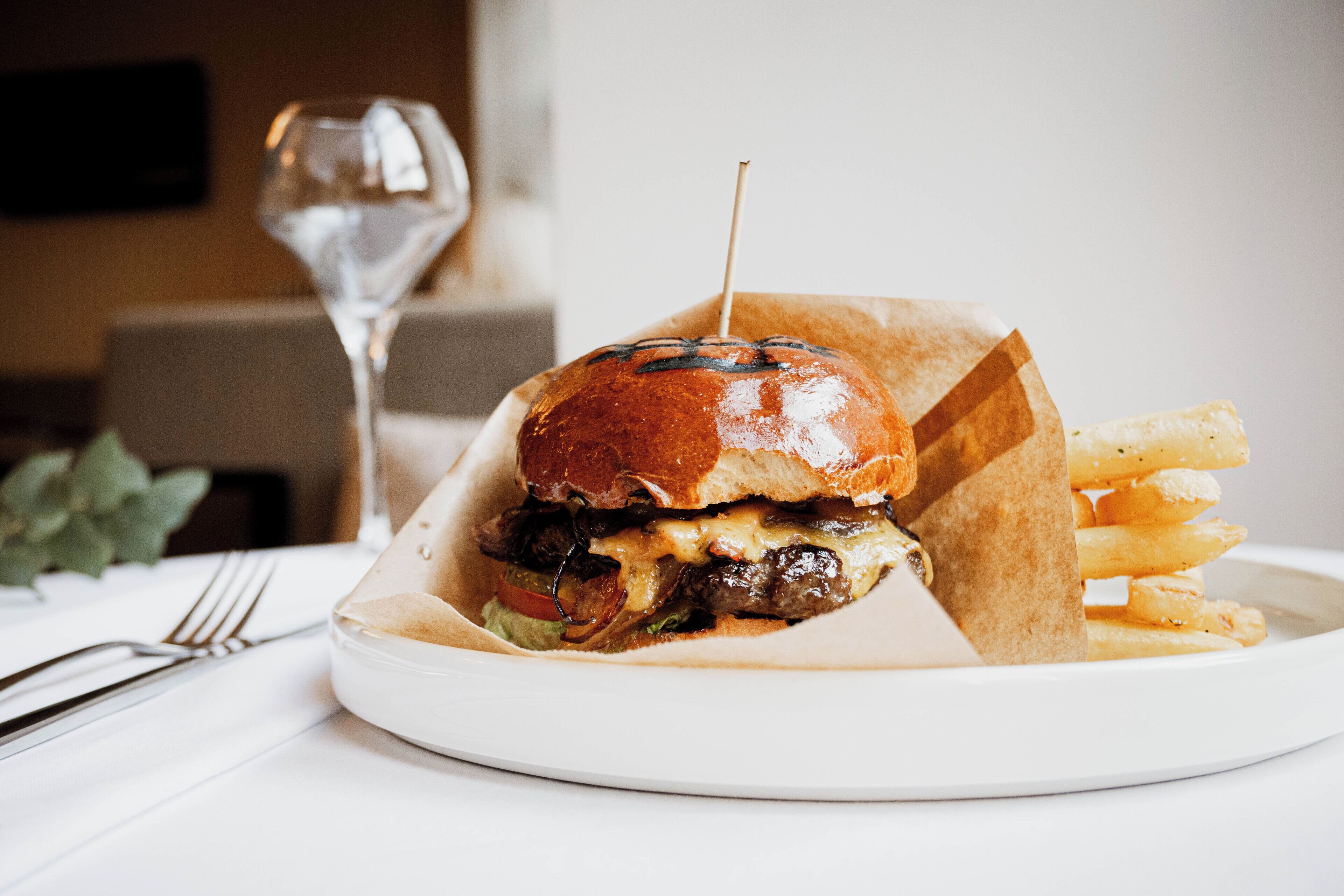 Ein Burger mit Pommes auf einem weißen Teller, ein Glas Wasser und eine Gabel auf einem Tisch.
