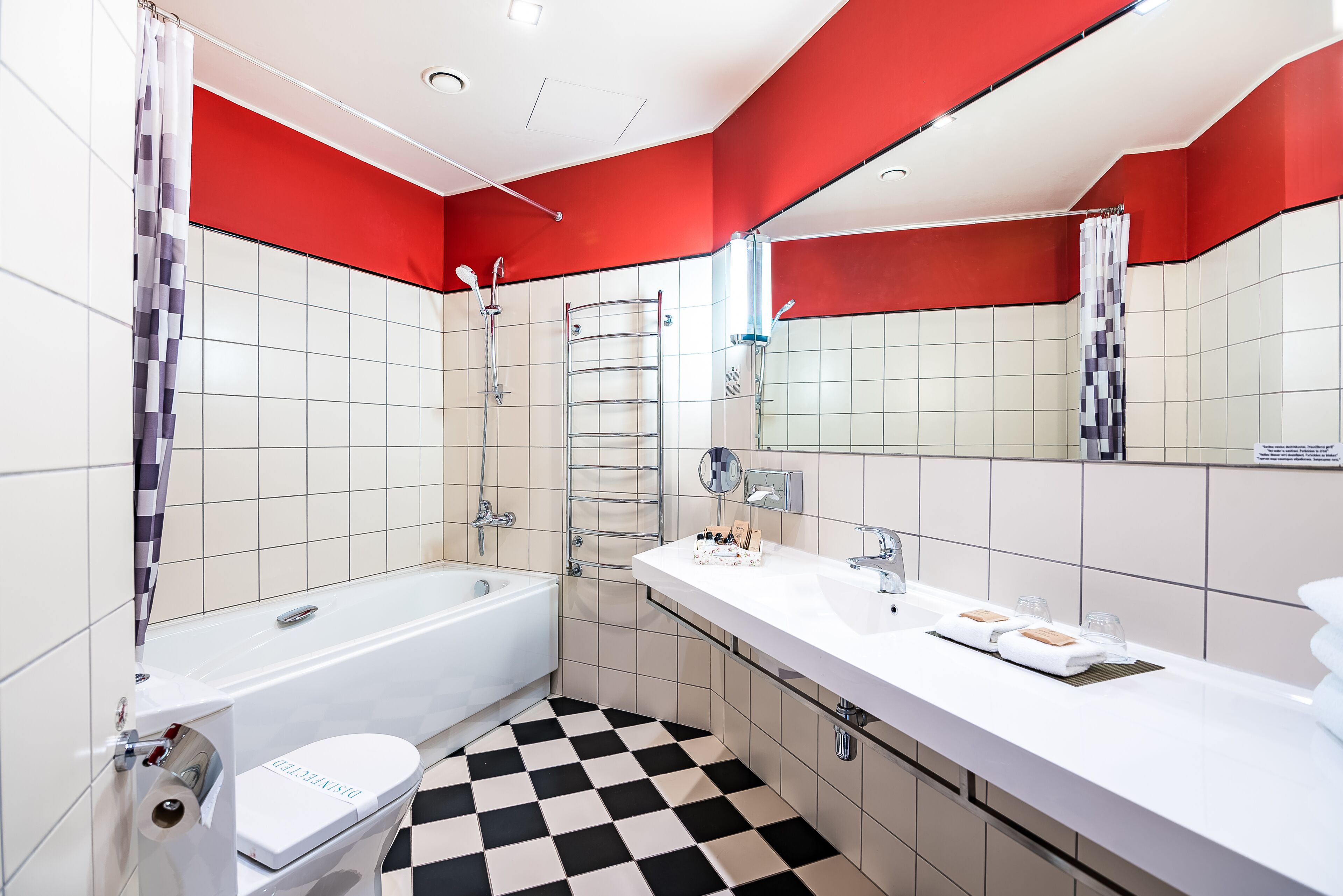 Un bagno moderno con pavimento a scacchi, pareti bianche e rosse, una vasca, una doccia e un mobile lavabo con asciugamani e articoli da toilette.
