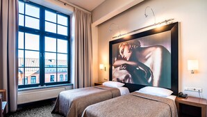 Habitación de hotel con dos camas, un ventanal con cortinas, un cuadro en la pared y una mesita de noche con lámpara.