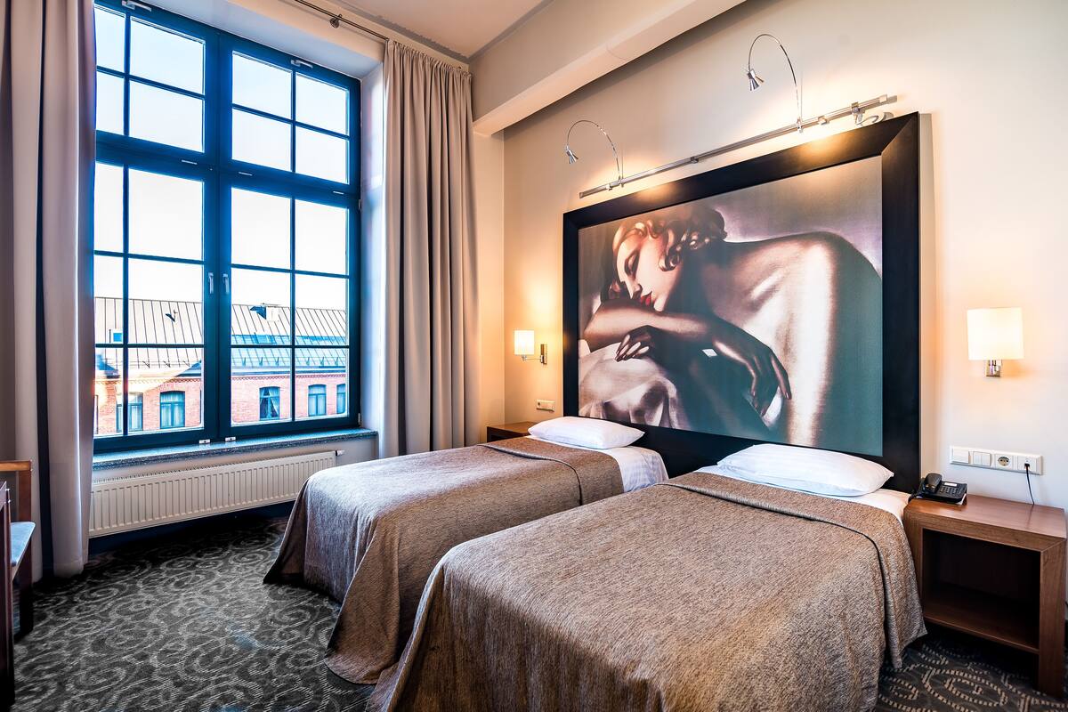 A hotel room with two beds, a large window with curtains, a painting on the wall, and a bedside table with a lamp.