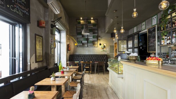 A cozy cafe interior with wooden tables and chairs, a bar counter with a display of snacks, and a chalkboard menu. - Hotel Robinson (Rome)