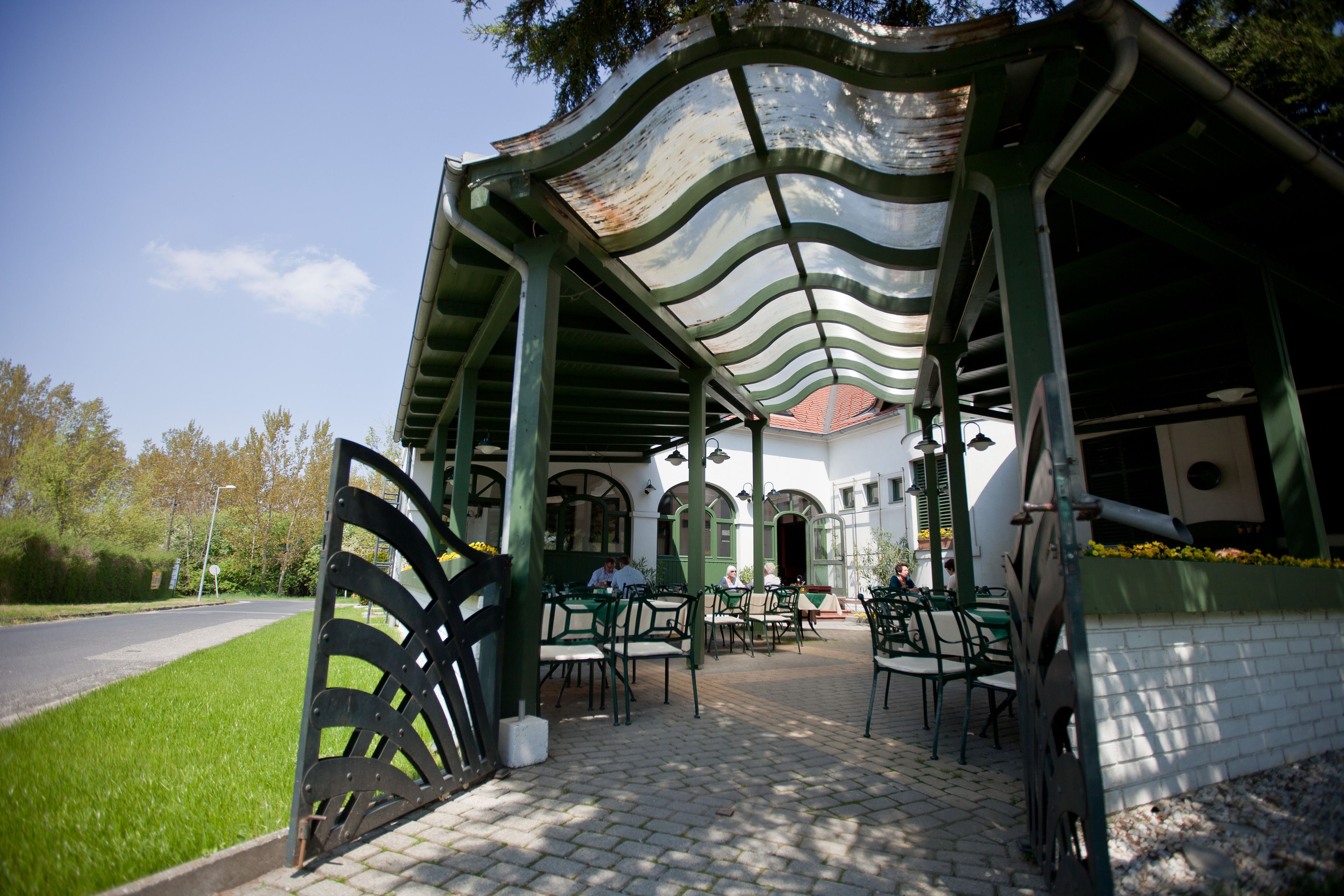 Ein Café im Freien mit gewölbtem Dach, grünen Stühlen und Tischen vor einem weißen Gebäude mit Rundbogenfenstern.