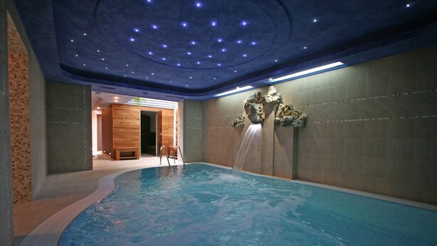 An indoor swimming pool with a waterfall feature, surrounded by tiled walls and a wooden bench. - Tornácos Panzió (HEGYKÖ)