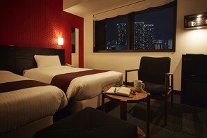 Une chambre d’hôtel avec deux lits, un bureau avec un carnet et une tasse, une chaise, et une vue sur le paysage urbain la nuit.