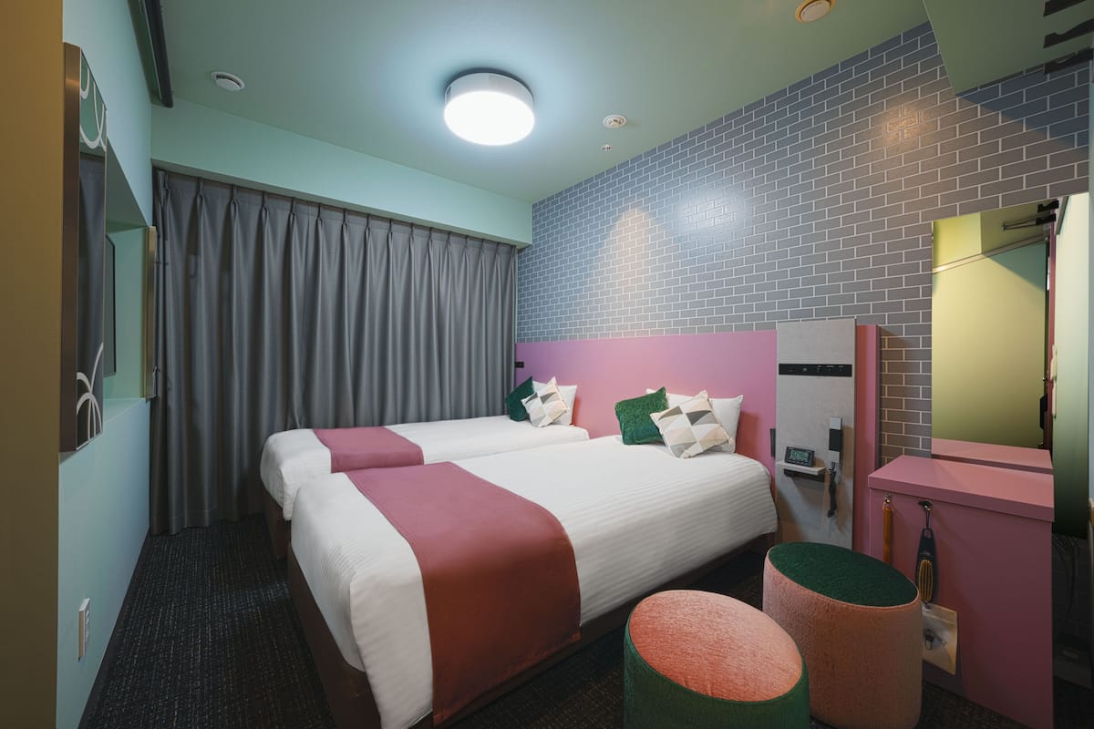 A hotel room with two beds, a large mirror, a vanity with a pink cabinet, and a green ottoman.