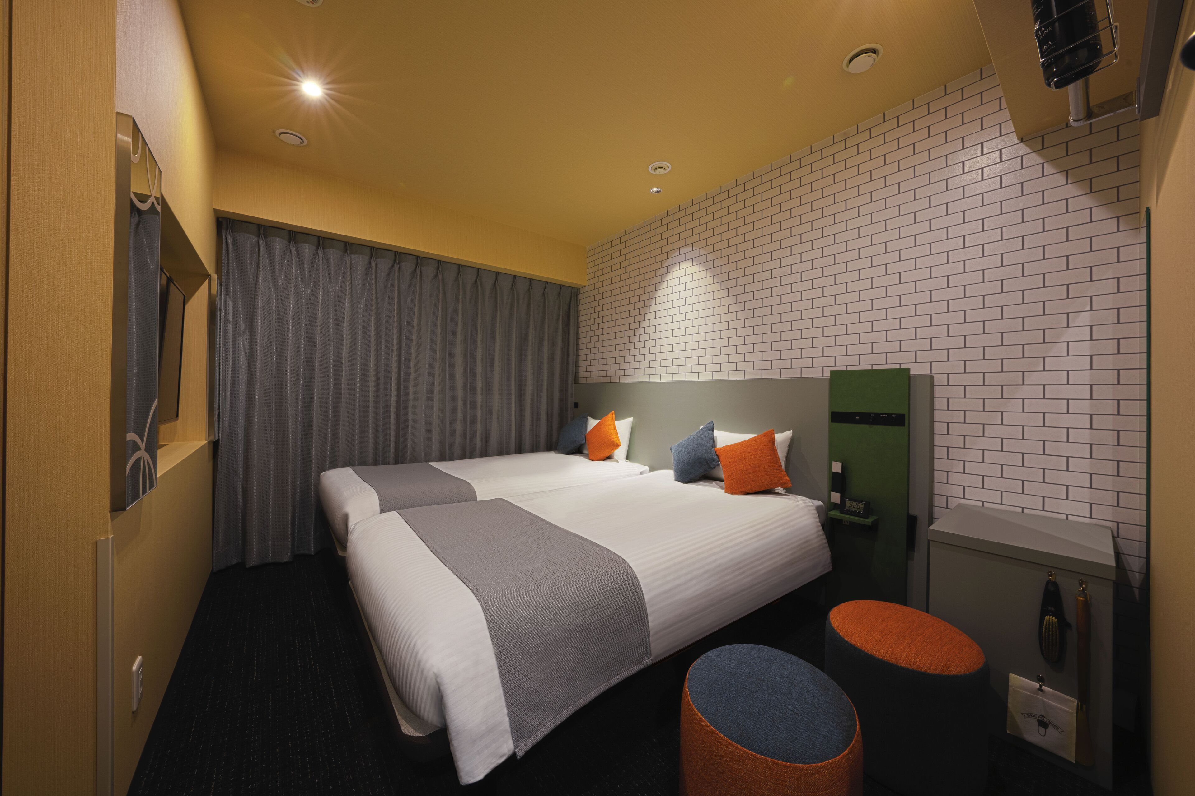 Quarto de hotel com duas camas, um banquinho redondo laranja, uma mesa de cabeceira verde e uma parede de tijolos brancos.