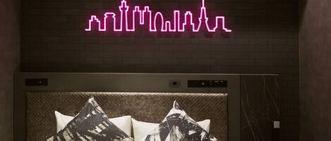 A hotel room with a neon city skyline sign, a bed with patterned pillows, and a marble headboard.