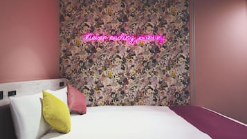 A hotel room with a floral wallpaper, a bed with white bedding and colorful pillows, and a neon sign that reads "Never-ending journey".