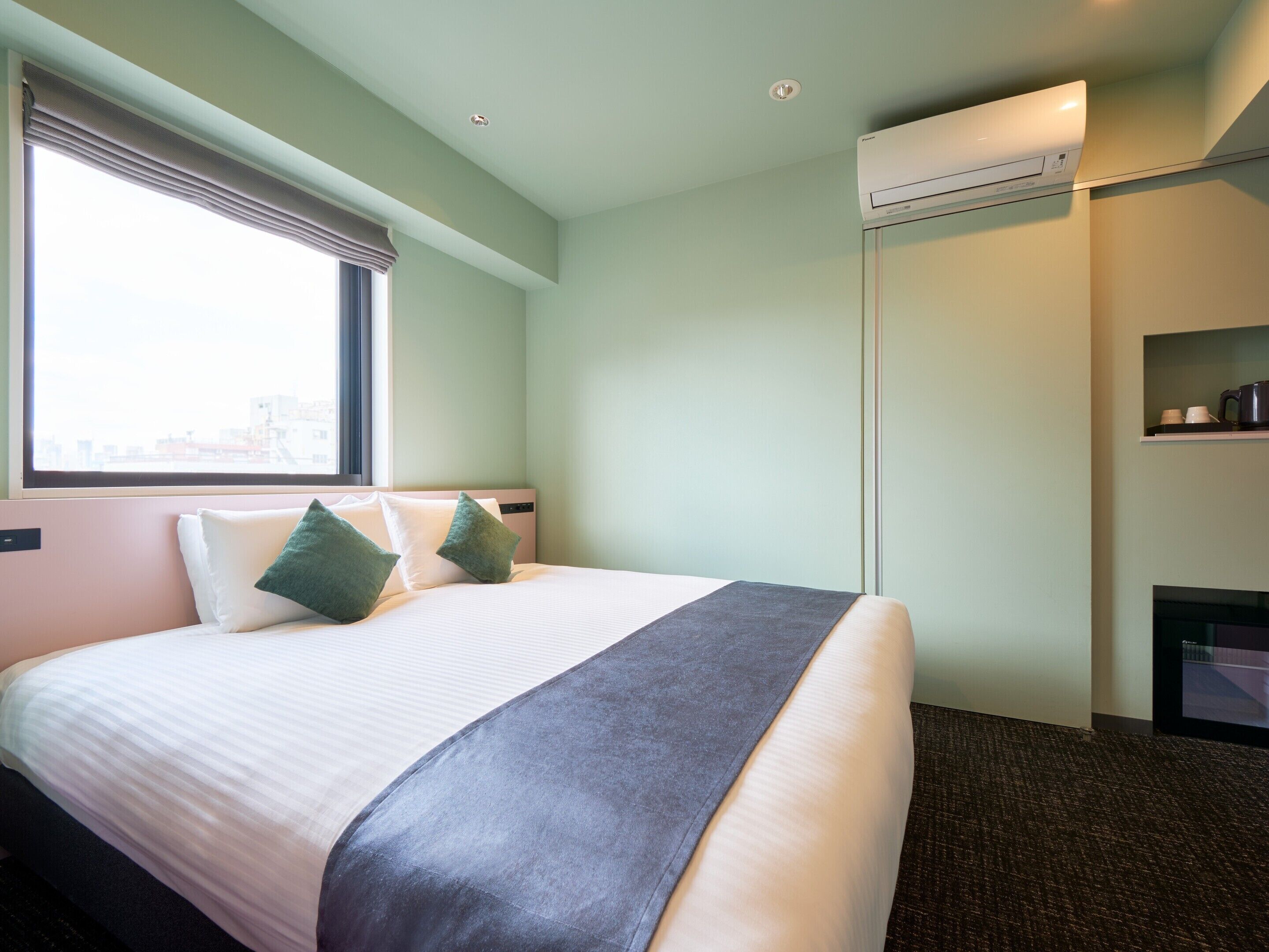 Habitación de hotel con cama, ventana con persianas, aire acondicionado integrado y un estante con objetos.