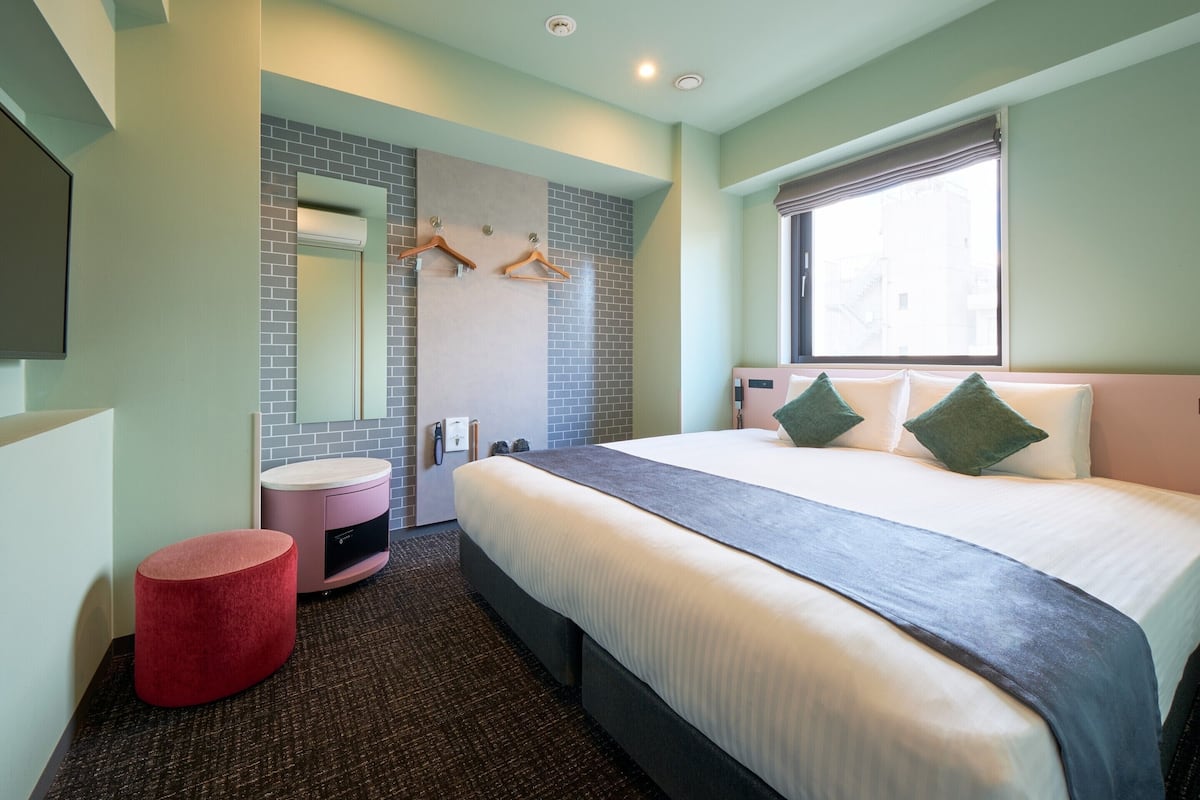 A hotel room with a large bed, a round red ottoman, a bathroom with a mirror and towel rack, and a window with a view.