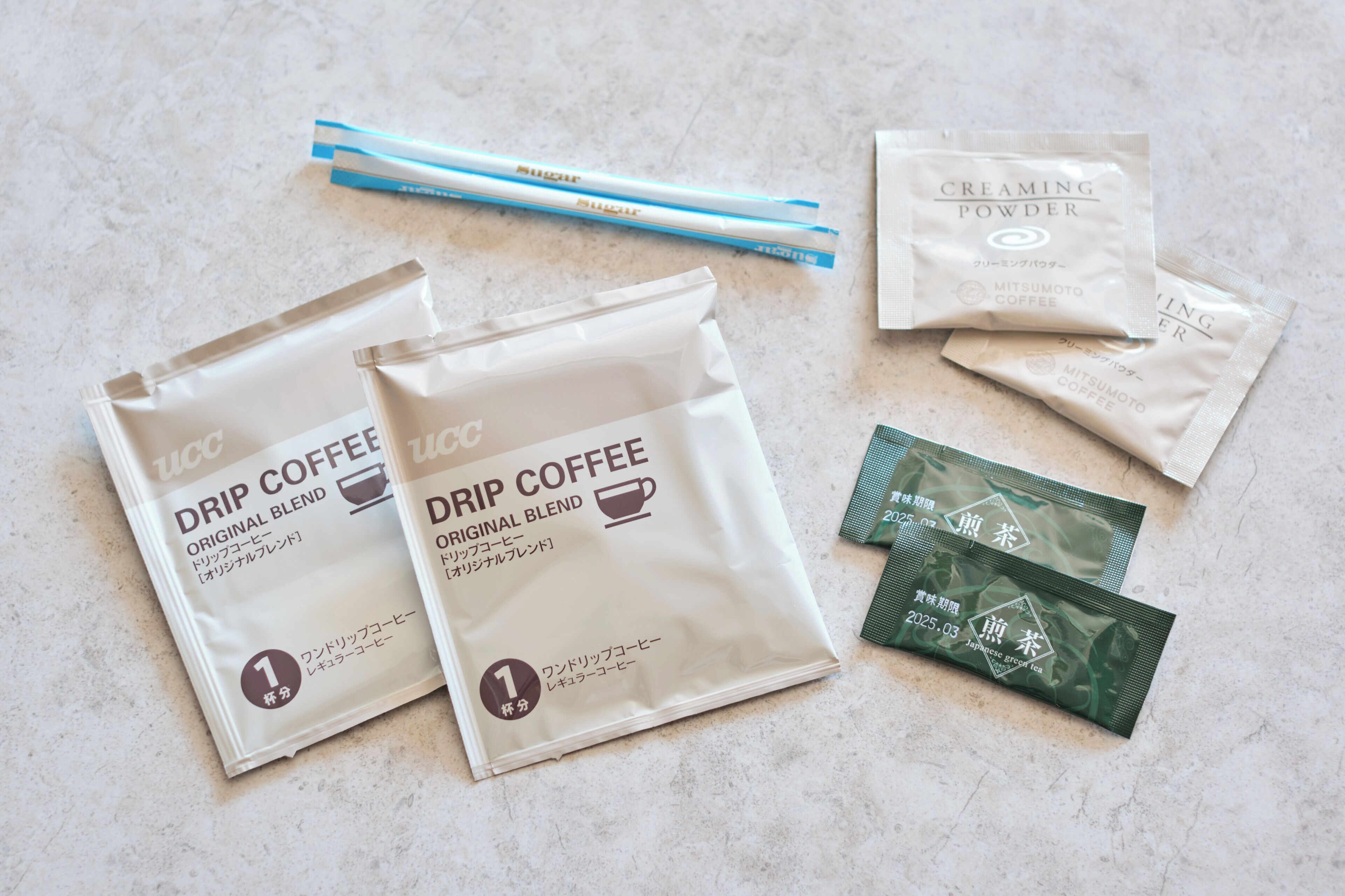 Two bags of drip coffee and two packets of creaming powder on a textured surface.