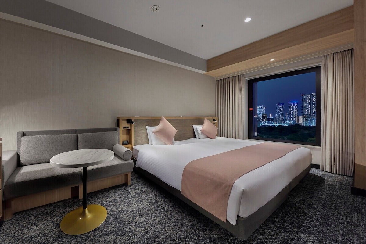 Quarto de hotel com cama, sofá, escrivaninha e vista para a cidade à noite.