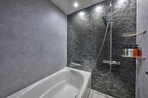 A modern bathroom with a white bathtub, a shower with a handheld showerhead, and a glass shelf with toiletries.