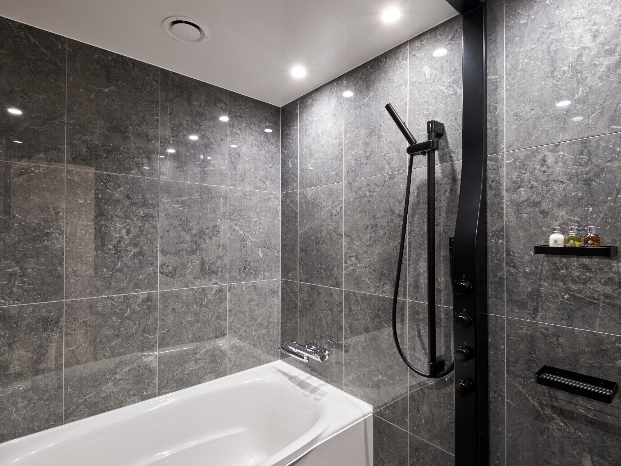 A modern bathroom with a white bathtub, dark marble tiles, a black shower system, and a small shelf with toiletries.