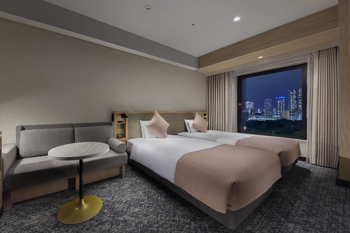 Quarto de hotel com cama, sofá, escrivaninha e vista para a paisagem urbana à noite.