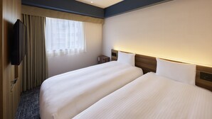 Hotellihuone, jossa on kaksi sänkyä, televisio, ikkuna, jossa on verhot, ja sängynpääty, jossa on sisäänrakennettu valaistus.