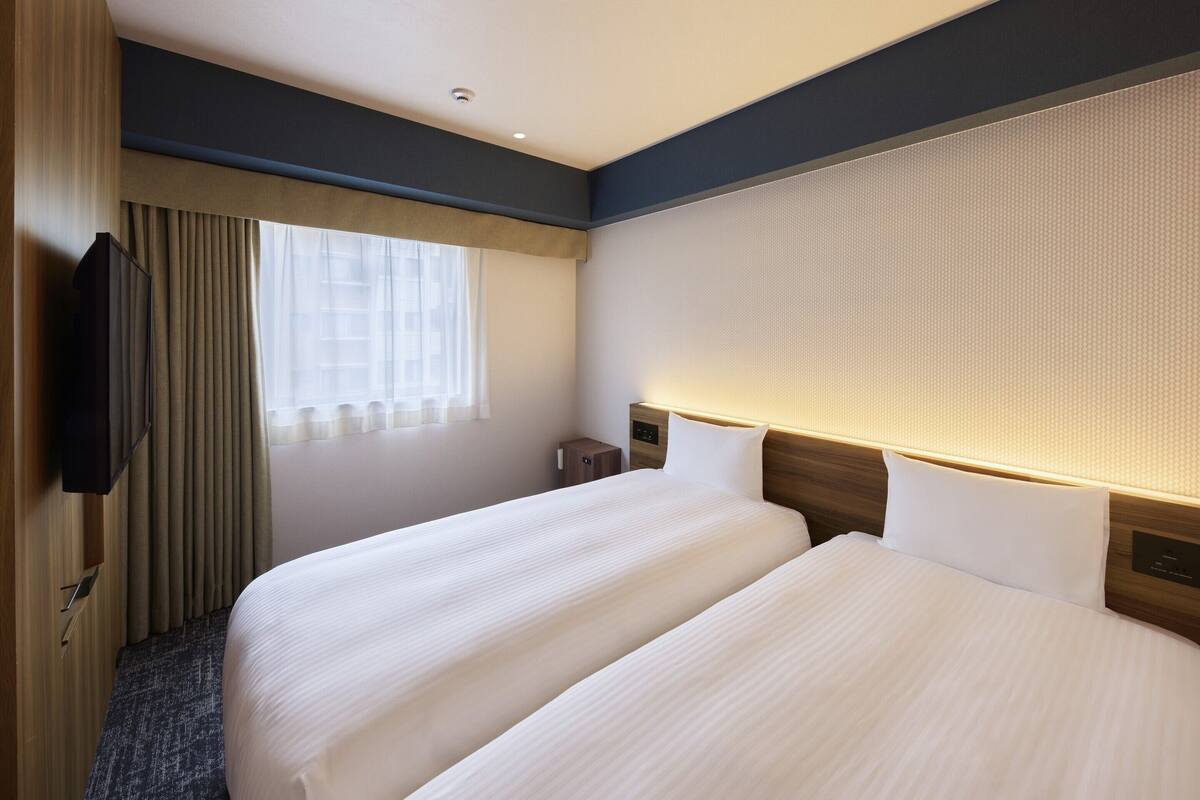 Ein Hotelzimmer mit zwei Betten, einem Fernseher, einem Fenster mit Vorhängen und einem Kopfteil mit integrierter Beleuchtung.