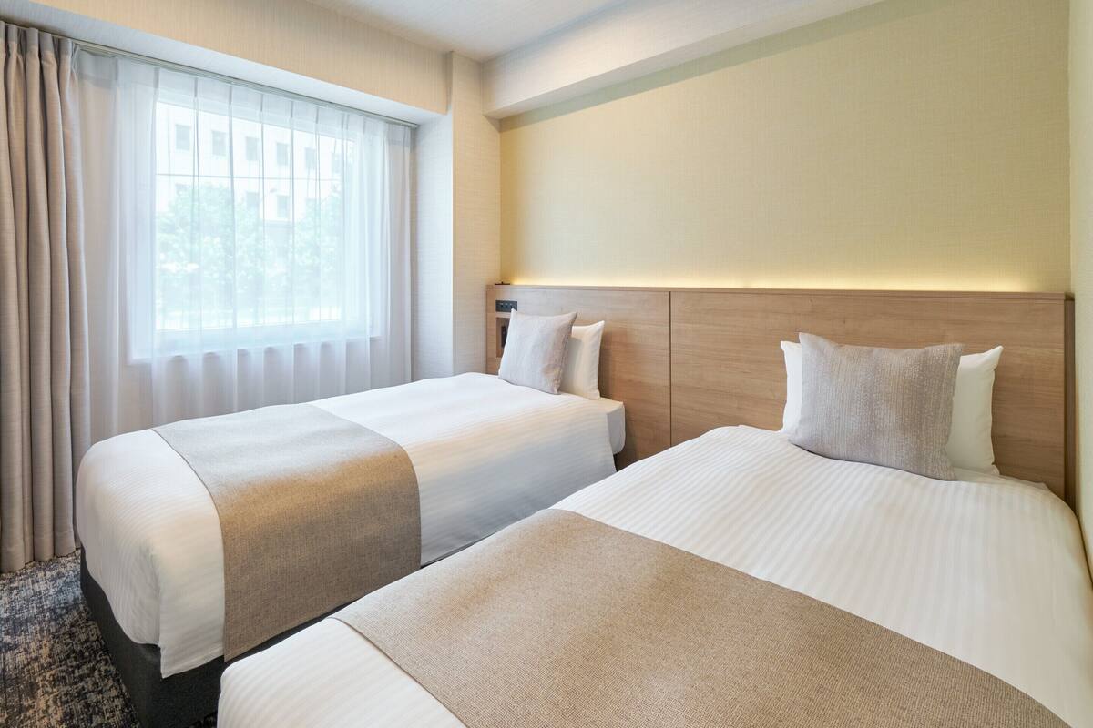 A hotel room with two beds, a large window with curtains, a wooden headboard, and a wall-mounted light.