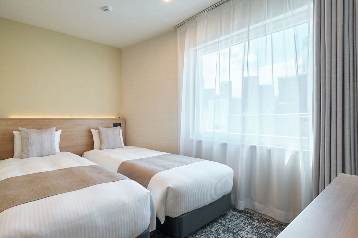 A hotel room with two beds, a headboard with a light fixture, a large window with sheer curtains, and a carpeted floor.