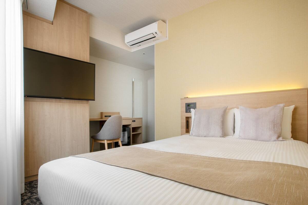 A hotel room with a bed, a desk, a chair, an air conditioning unit, and a television.