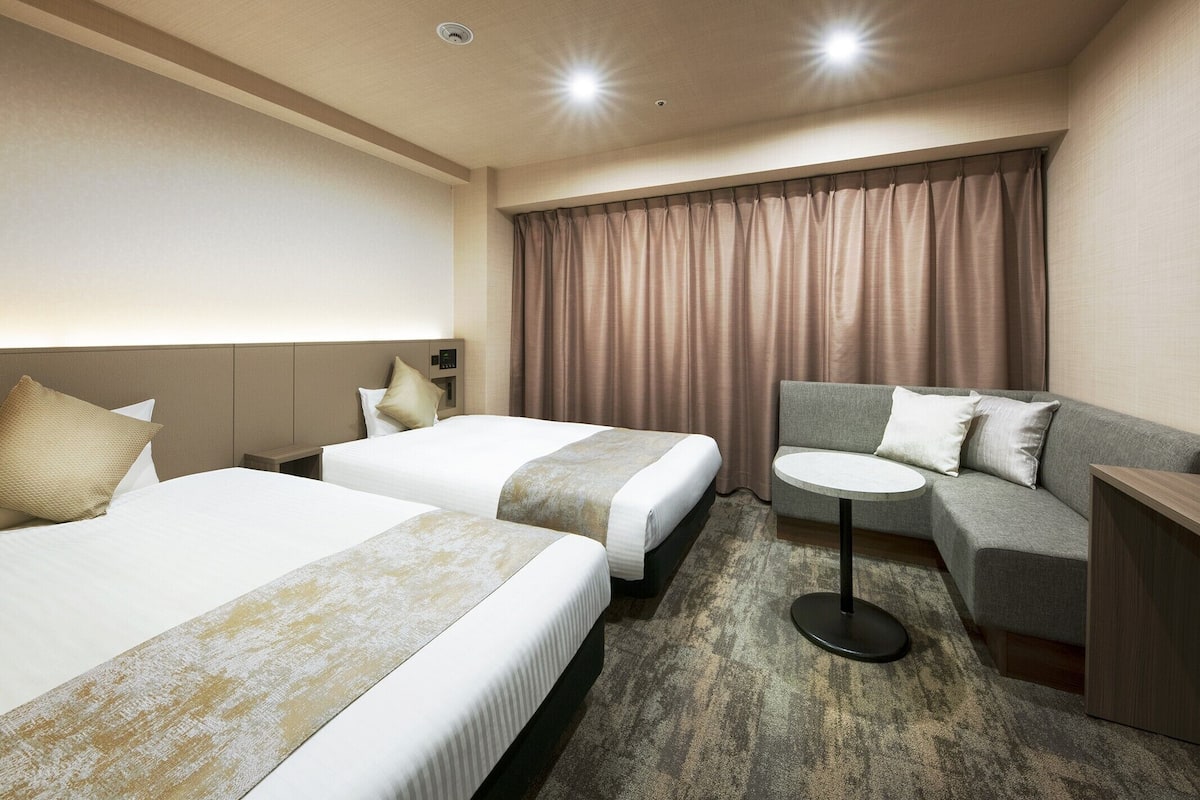 A hotel room with two beds, a sofa, a small table, and a headboard with a control panel.