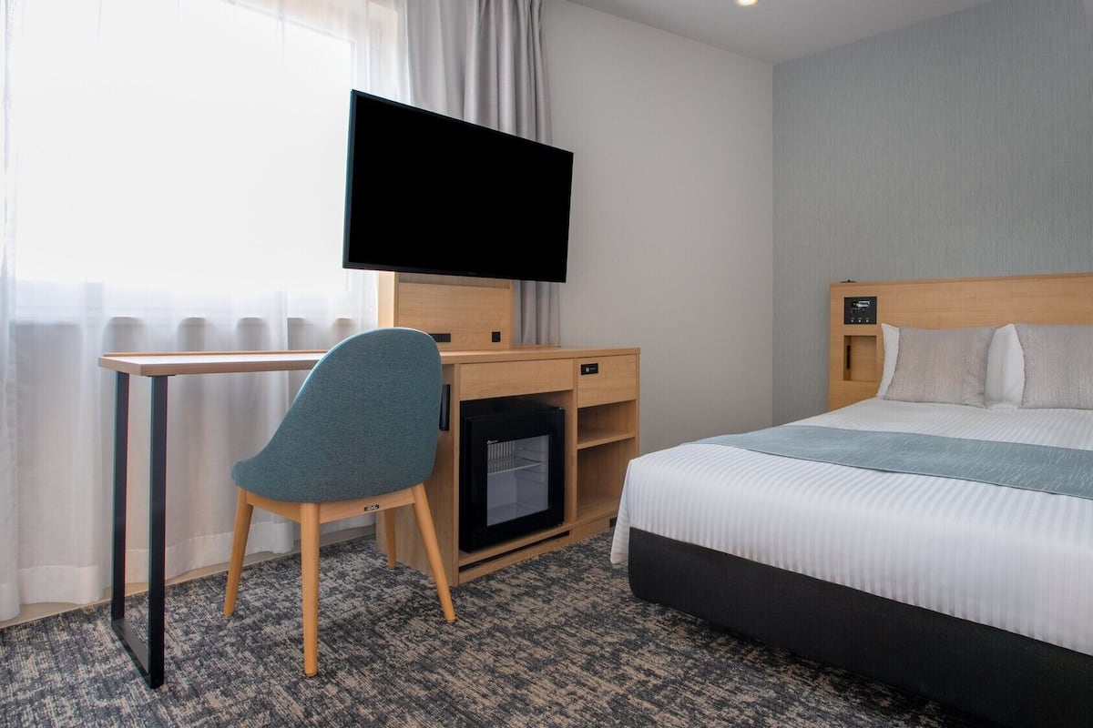 Una habitación de hotel con cama, escritorio, silla, televisor y un pequeño calentador.