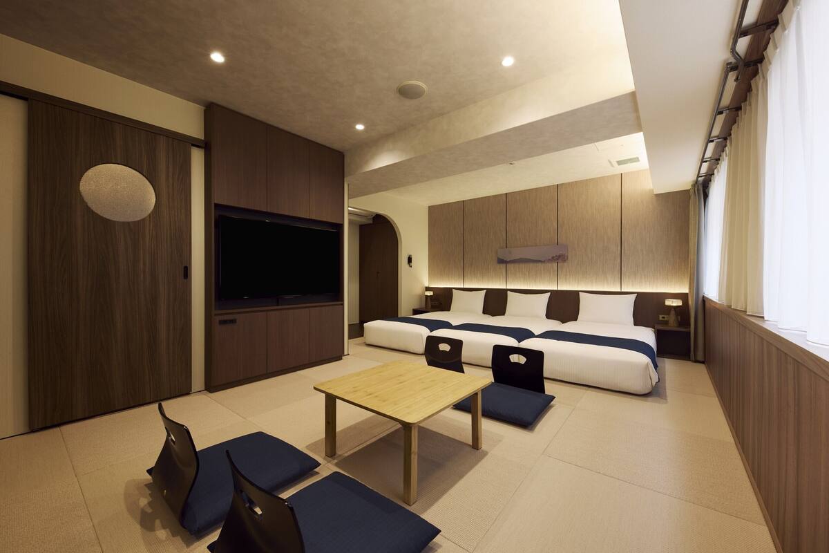 A modern hotel room with a large bed, a flat-screen TV, a wooden table, and a seating area with cushions.