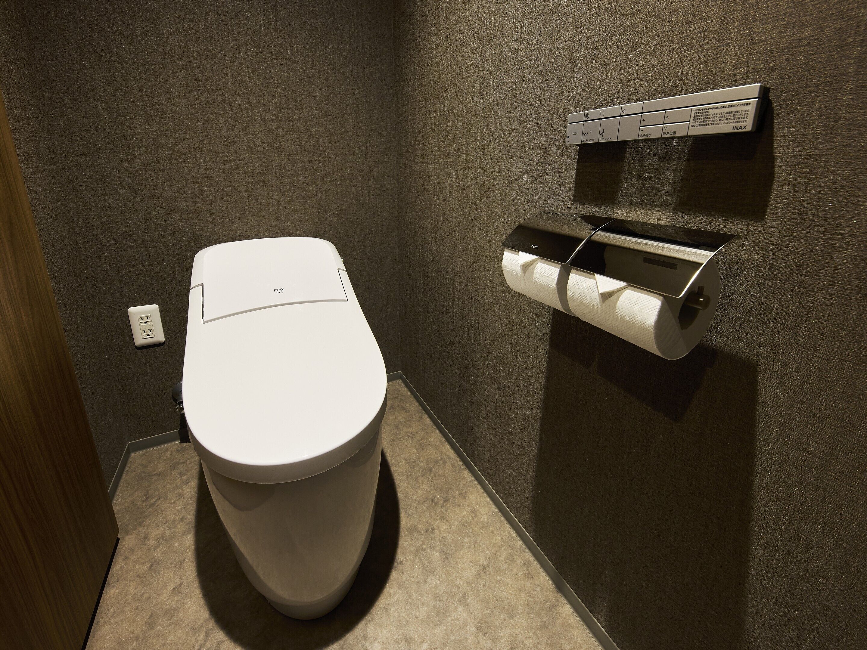 Un inodoro moderno con un dispensador de toallas de papel en la pared, un enchufe y una pared con textura.