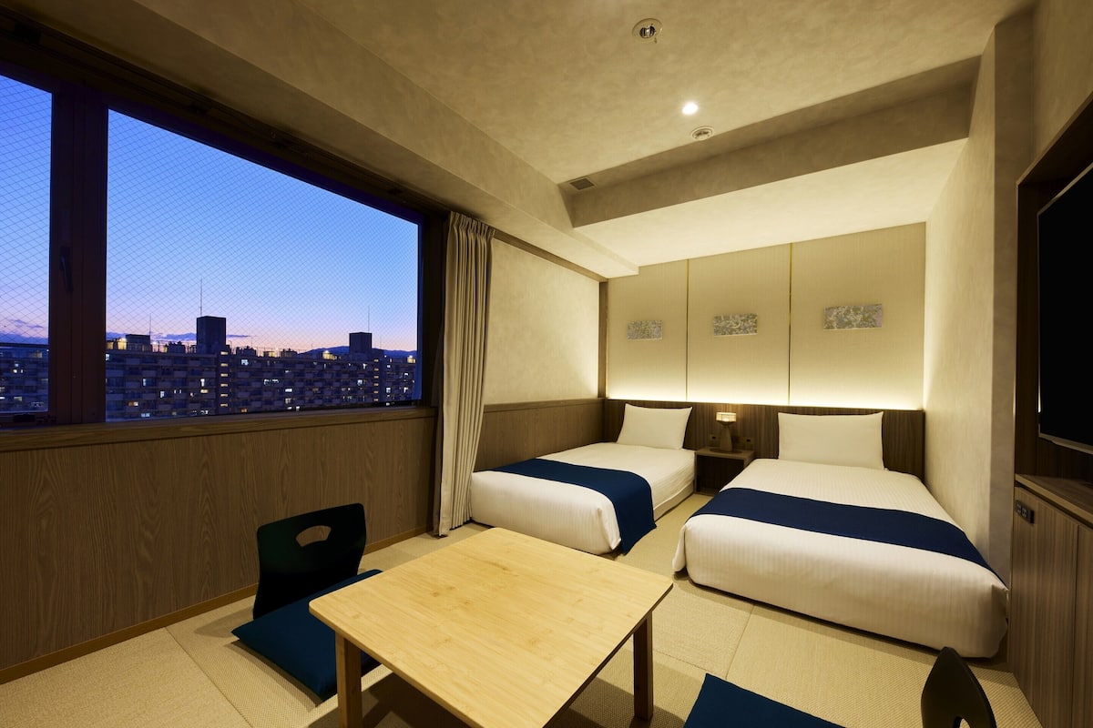 A hotel room with two beds, a small table, and a view of a cityscape at dusk.